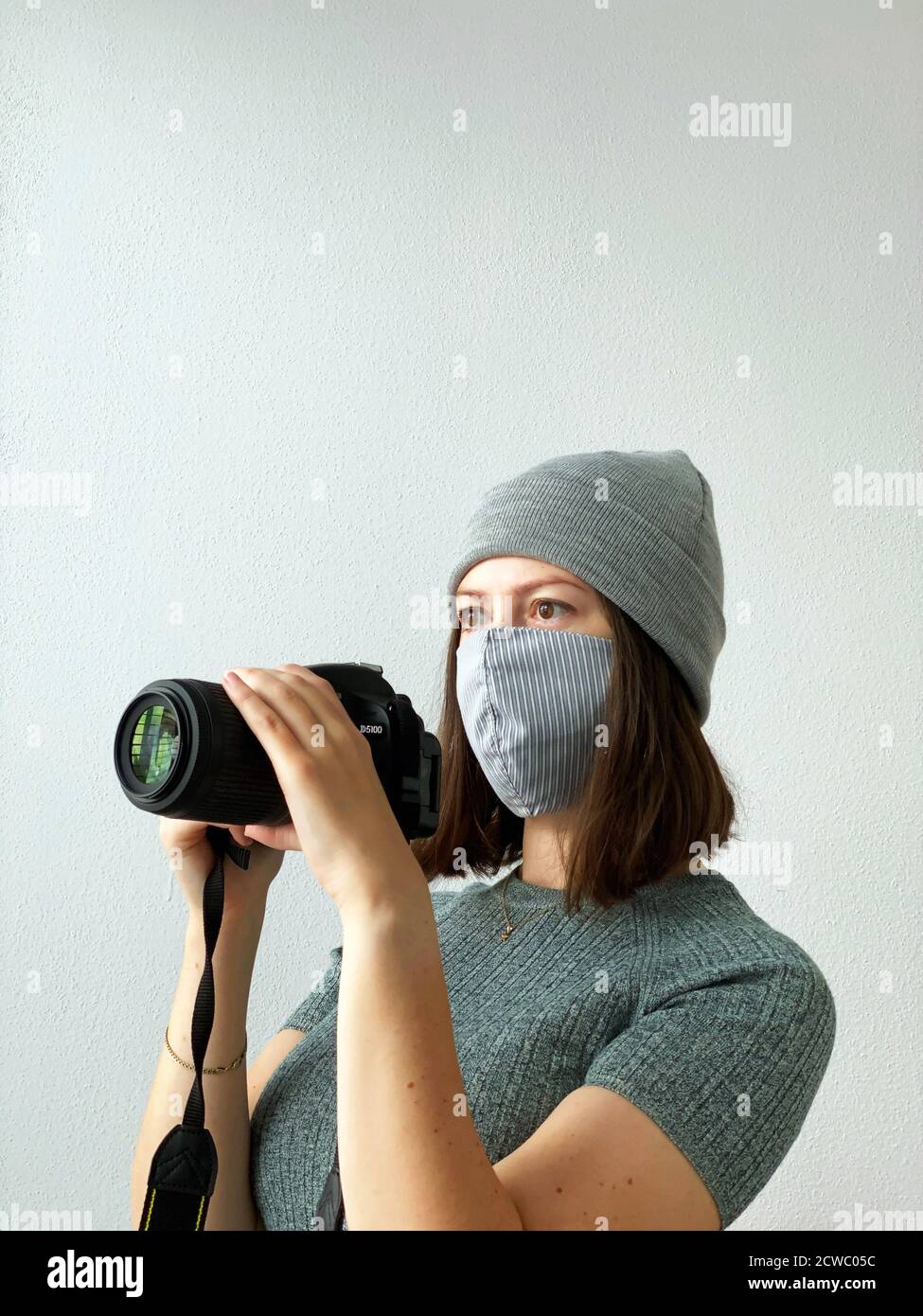 Schießen in der Zeit der Pandemie. Junge Fotografin mit Kamera in einer wiederverwendbaren Stoffmaske und grauem Hut und grünem Pullover mit kurzen braunen Haaren Stockfoto