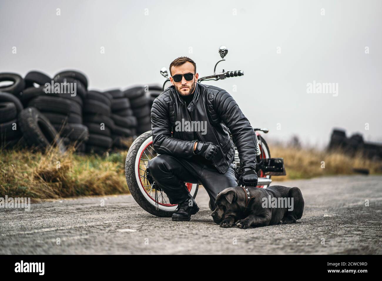 Biker in einem Lederanzug kauerte neben seinem Hund und rotem Motorrad auf der Straße. Viele Reifen im Hintergrund. Stockfoto