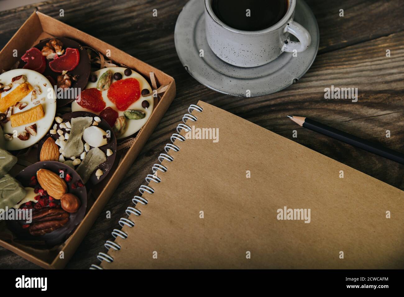 Pralinen mit getrockneten Früchten in einer Schachtel und Bastelpapier-Notizbuch in der Nähe der Tasse Kaffee. Ort für Ihren Text. Stockfoto