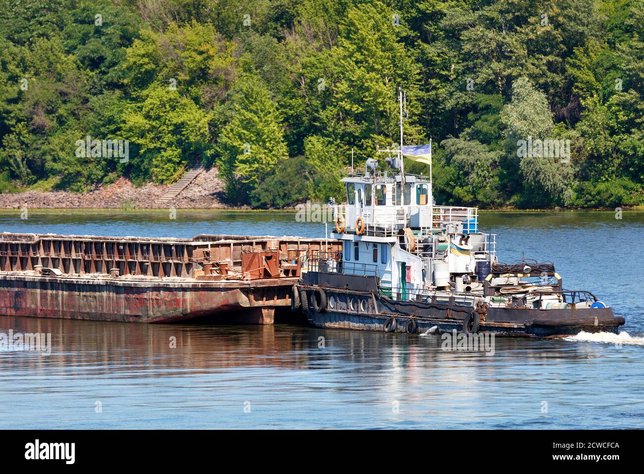 Flussschlepper langsam schieben eine leere rostigen Barge den Fluss hinunter gegen Küstengrün, Fluss Güterverkehr Konzept, Copy Space. Stockfoto