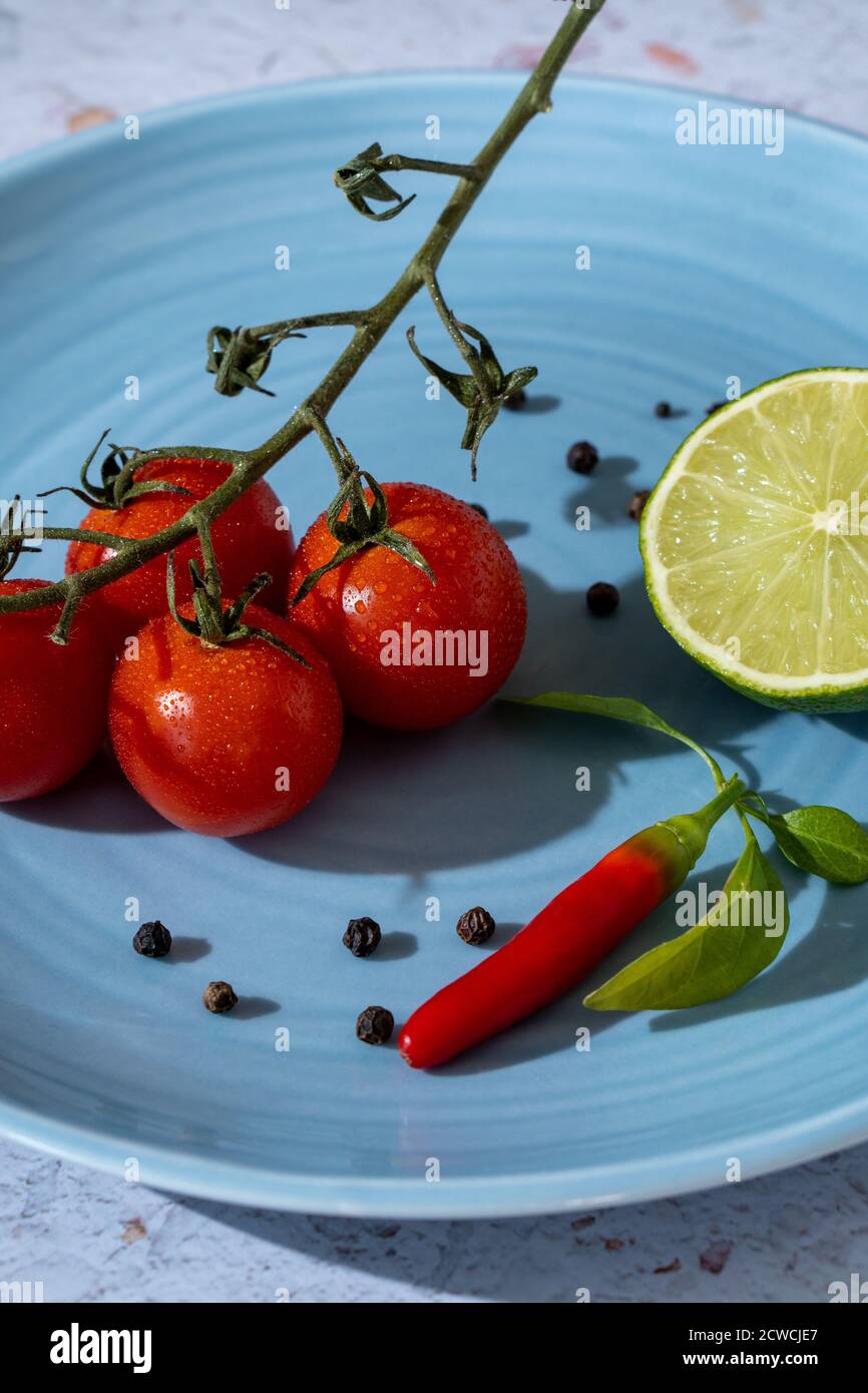 Minimalistisches Konzept für blutige Mary-Zutaten. Ein Zweig aus frischen und roten Kirschtomaten, schwarzen Pfeffersamen, einer Zitrone und einem würzigen Chili auf einem hellblauen Stockfoto