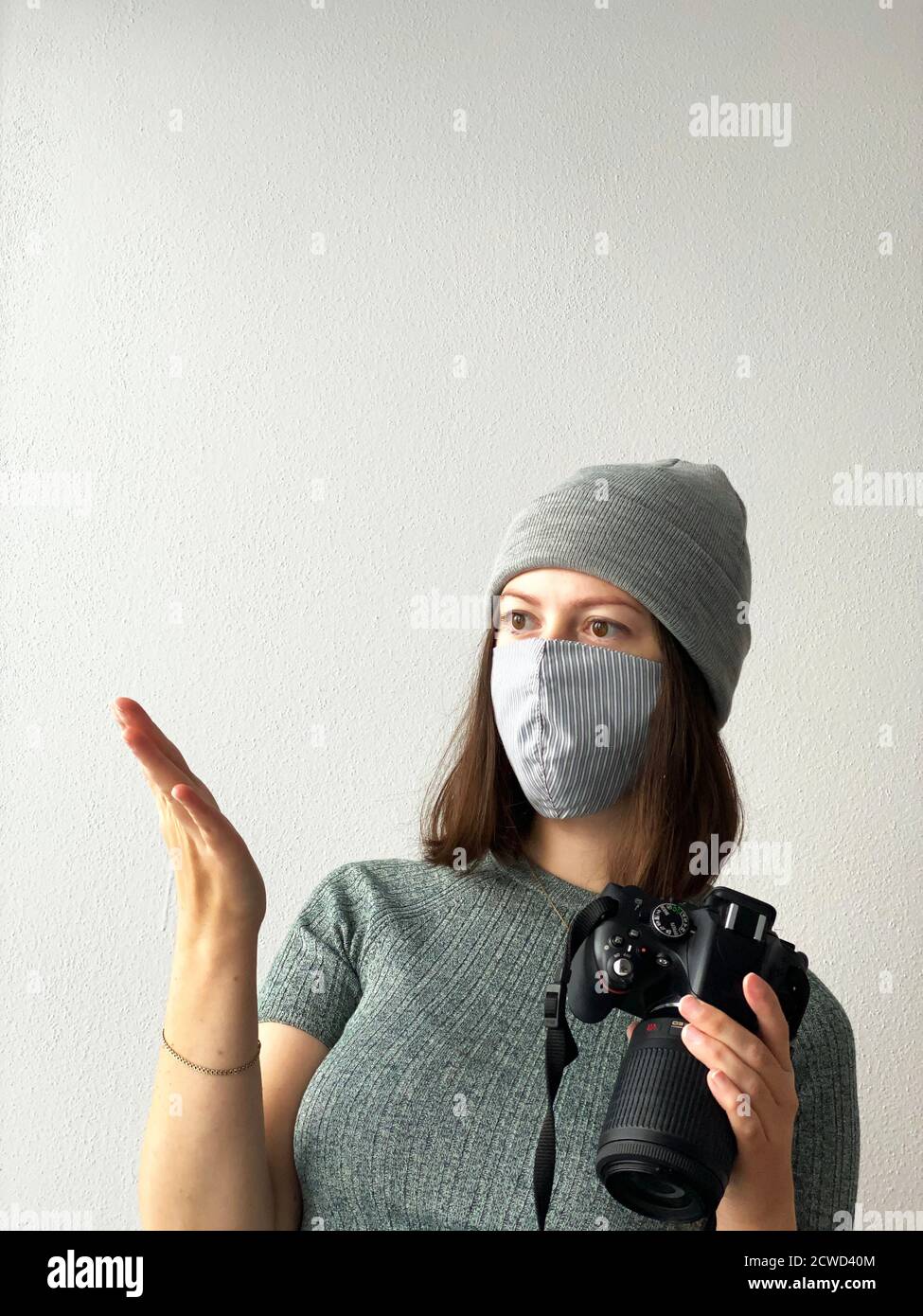 Schießen in der Zeit der Pandemie. Junge Fotografin mit Kamera in einer wiederverwendbaren Stoffmaske und grauem Hut und grünem Pullover mit kurzen braunen Haaren Stockfoto