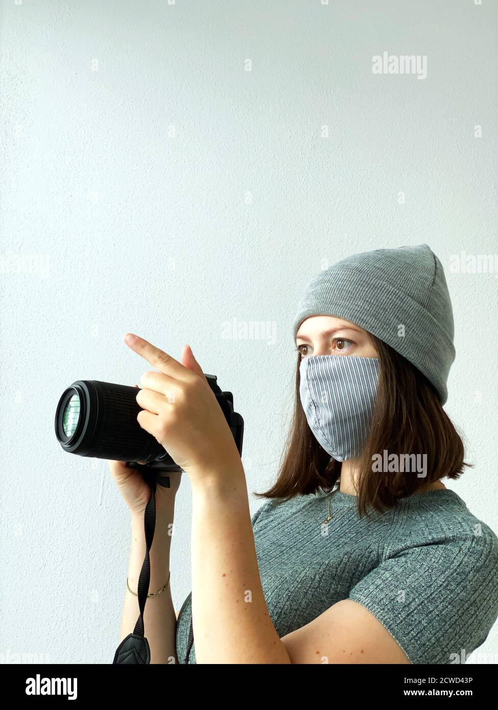 Schießen in der Zeit der Pandemie. Junge Fotografin mit Kamera in einer wiederverwendbaren Stoffmaske und grauem Hut und grünem Pullover mit kurzen braunen Haaren Stockfoto