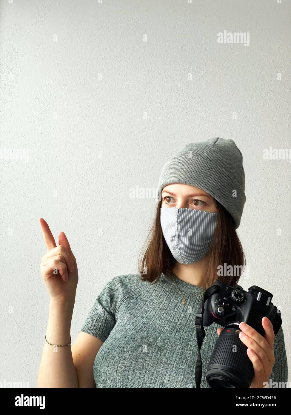 Schießen in der Zeit der Pandemie. Junge Fotografin mit Kamera in einer wiederverwendbaren Stoffmaske und grauem Hut und grünem Pullover mit kurzen braunen Haaren Stockfoto