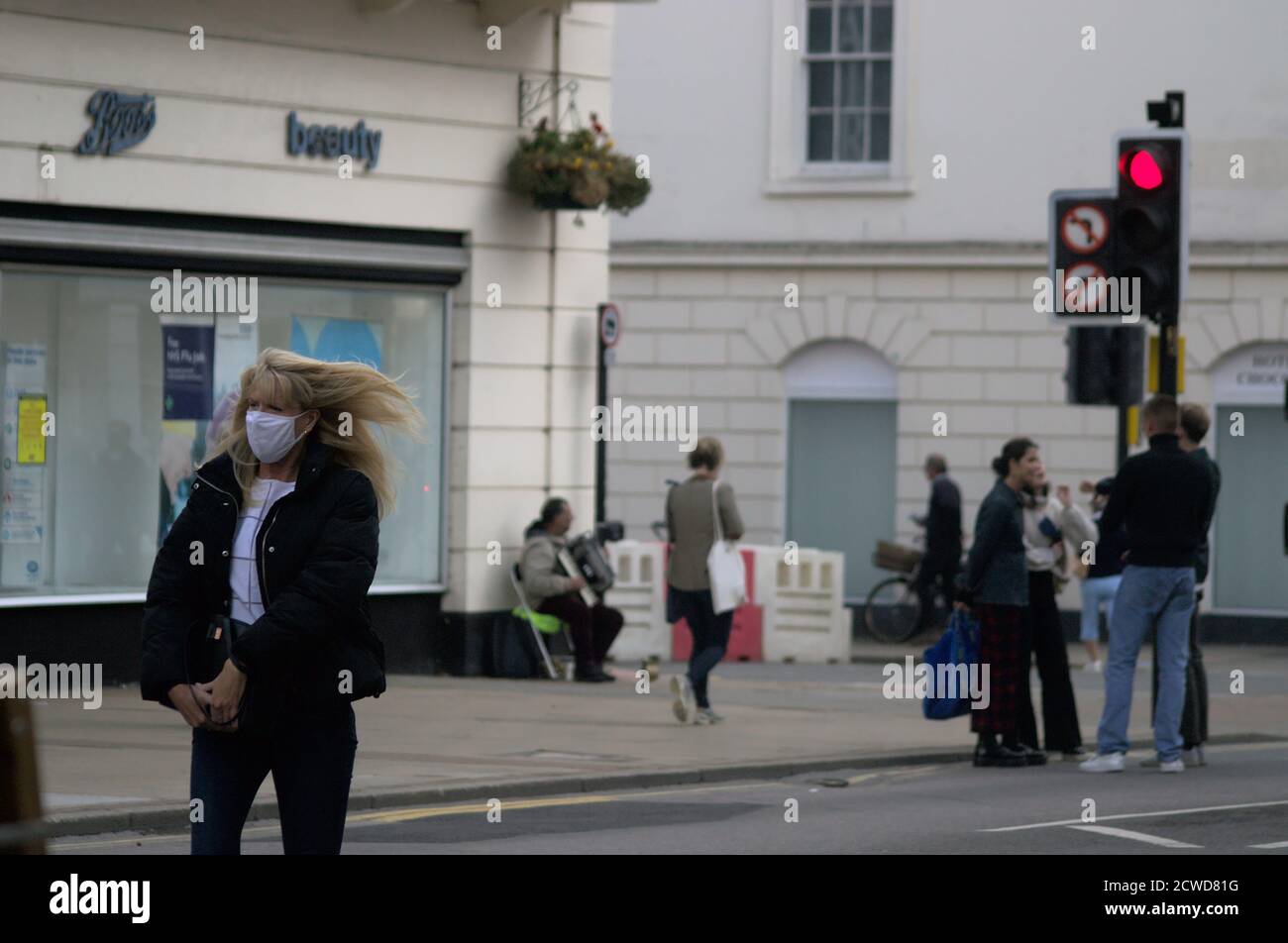 Eine Frau geht entlang der Parade in Leamington mit einer Gesichtsmaske, ihre Haare wehen im Wind. Im Hintergrund spielt ein Busker ein Akkordeon Stockfoto