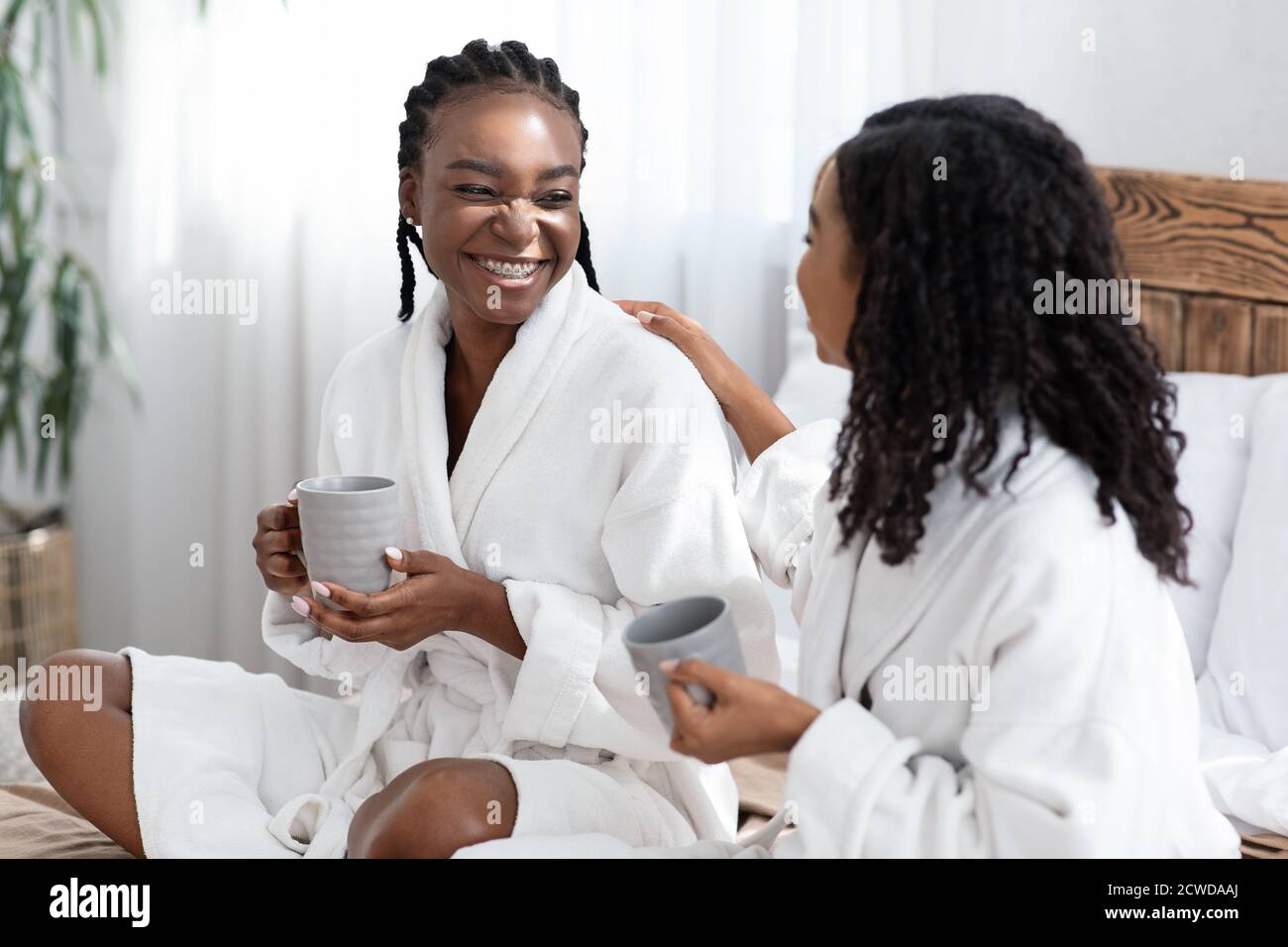 Zwei junge Frauen in Bademänteln sitzen auf dem Bett und trinken Kaffee Stockfoto