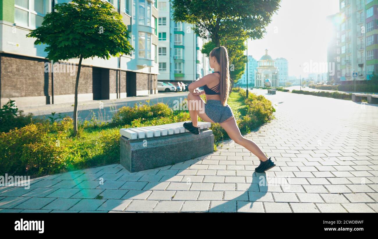 Unkenntlich Fitness Frau Stretching Bein mit Bank, tun Flexibilität Übungen nach dem Laufen Training in der Stadt Straßen. Junge Frau, die sich vor dem Training in Morgensonne aufwärmt. Stockfoto