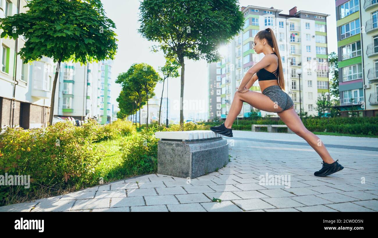 Muskulöse Frau in Sportswear Stretching Beine mit Bank, tun Flexibilität Übungen nach dem Training in der Stadt Straßen laufen. Flexible Bodybuilderin wärmt sich unter der Morgensonne beim Joggen auf. Stockfoto