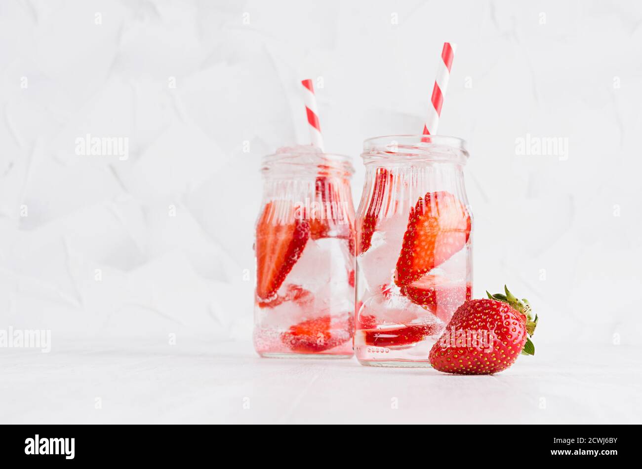 Sommer selbstgemachte Getränke - infundiertes Erdbeerwasser mit geschnittener Beere, Eiswürfeln, Soda, Stroh im weichen hellweißen Innenraum, Kopierraum. Stockfoto