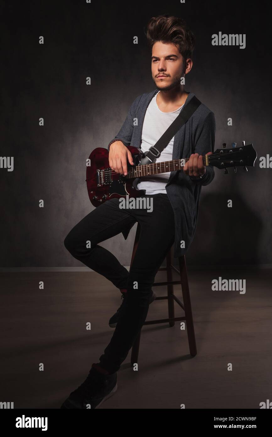 Skinny Rocker Gitarre spielen, während im Studio Hintergrund sitzen und Wegschauen Stockfoto