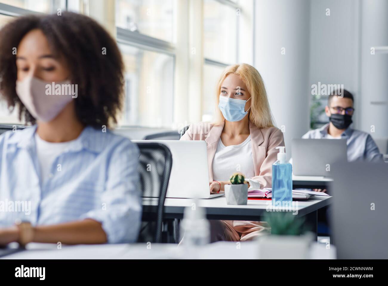 Arbeit der Mitarbeiter der Gesellschaft nach Coronavirus Quarantäne. Multirassische Arbeiter in Schutzmasken an Schreibtischen mit Laptops und Antiseptika Stockfoto