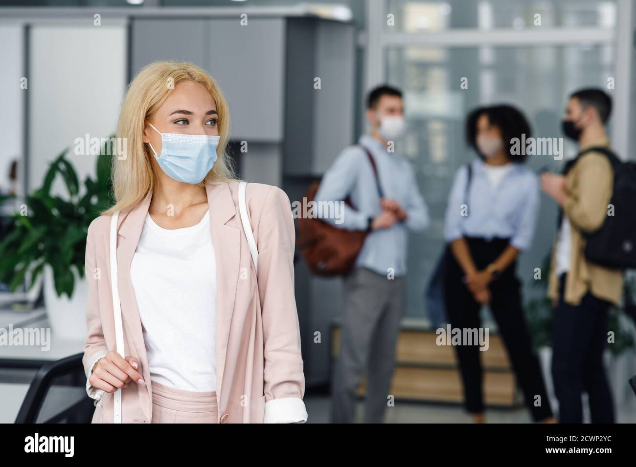 Nach Sperrung und neuer Normalität wieder zur Arbeit zurückkehren. Frau in Schutzmaske sieht zur Seite im Korridor des modernen Büros mit verschwommenen Kollegen Stockfoto