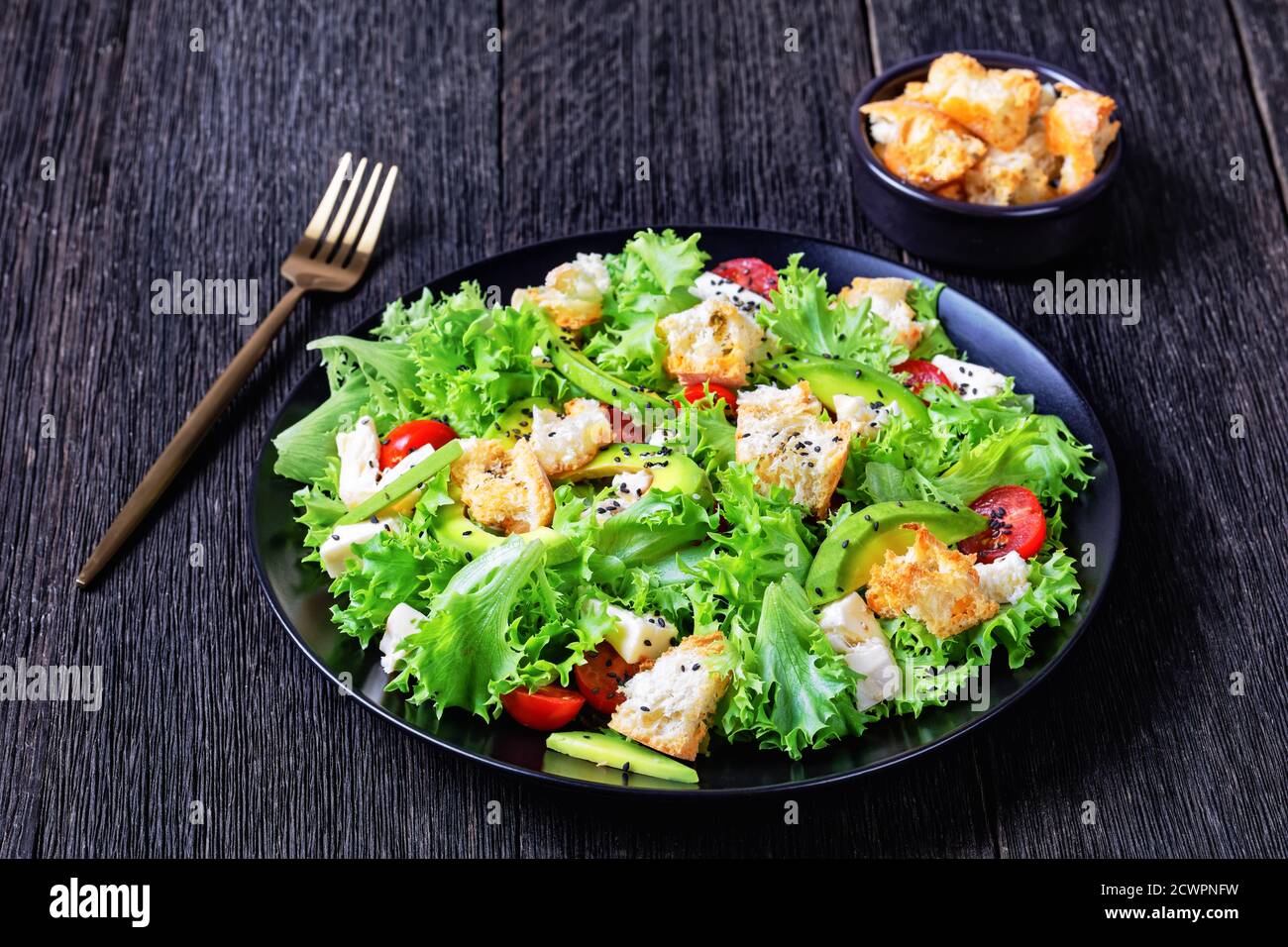 Blattsalat mit Avocado, Kirschtomaten, Mozzarella und Croutons bestreut mit schwarzen Sesamsamen auf einem Teller, auf einem Holztisch, Landschaftsblick Stockfoto