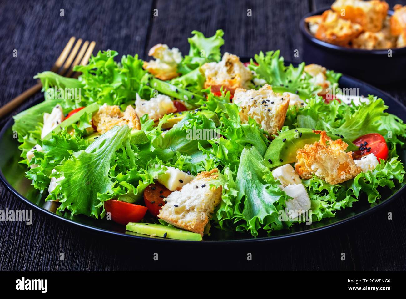 Blattsalat mit Avocado, Kirschtomaten, Mozzarella und Croutons bestreut mit schwarzen Sesamsamen auf einem Teller, auf einem Holztisch, Landschaftsblick Stockfoto