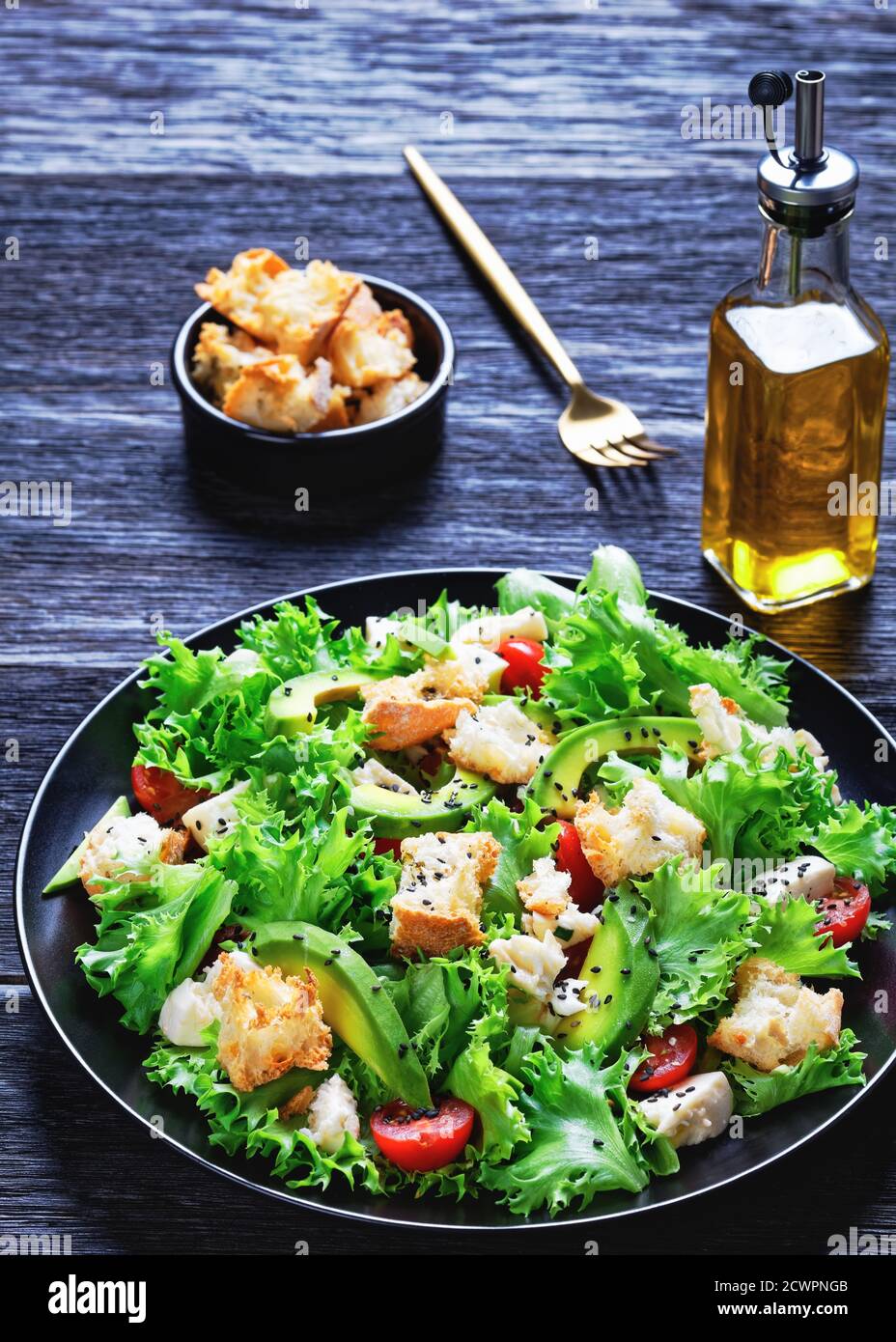 Blattsalat mit Avocado, Kirschtomaten, Mozzarella und Croutons bestreut mit schwarzen Sesamsamen auf einem Teller, auf einem Holztisch, vertikale Ansicht von Stockfoto