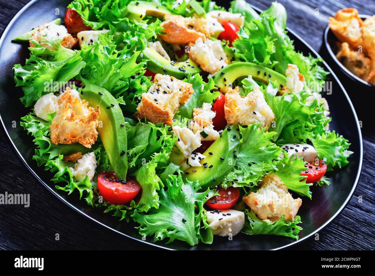 Blattsalat mit Avocado, Kirschtomaten, Mozzarella und Croutons bestreut mit schwarzen Sesamsamen auf einem Teller, auf einem Holztisch, Landschaftsblick Stockfoto