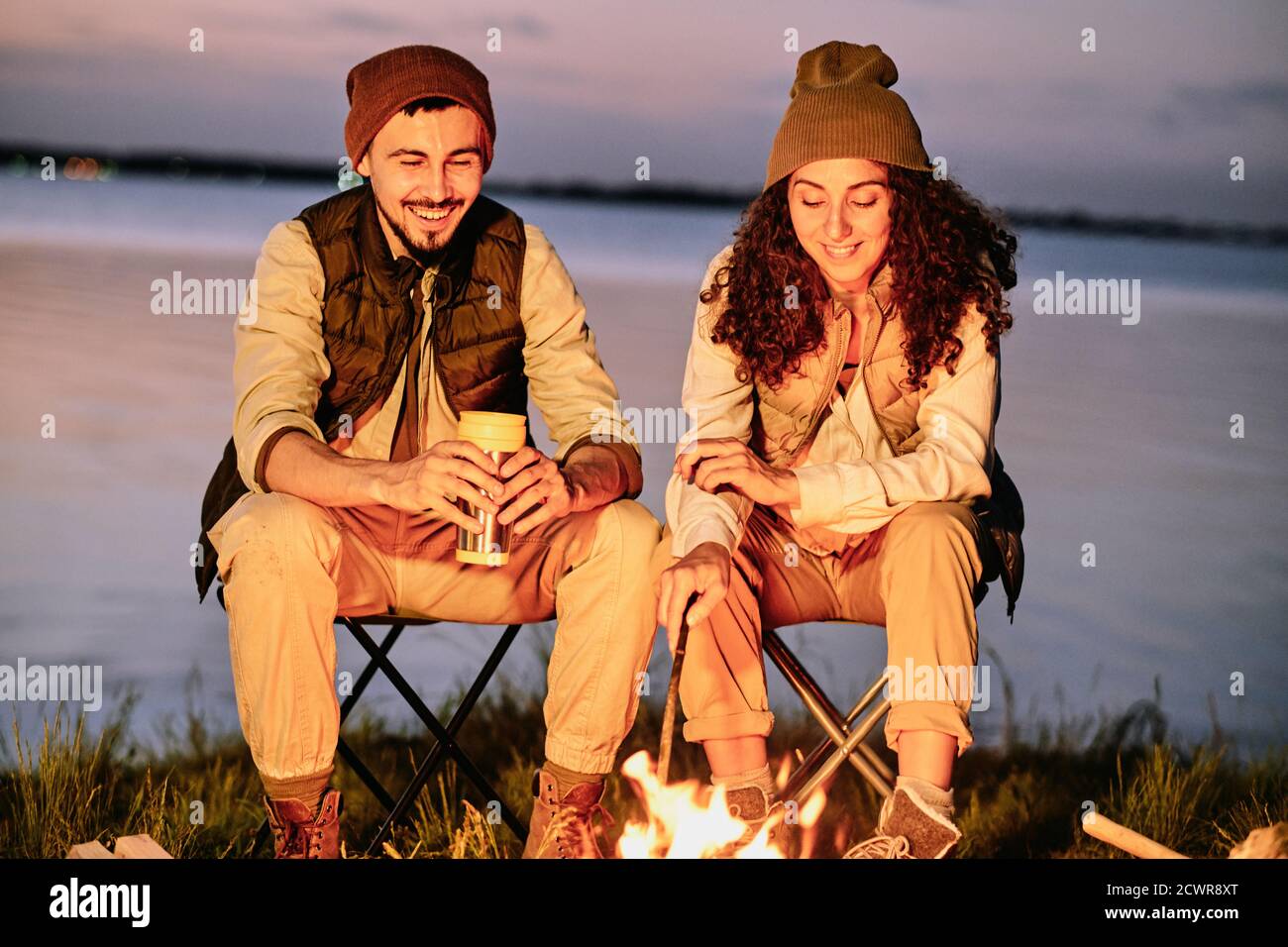 Fröhliches junges Paar in Hüten sitzen auf Klappstühlen in der Nähe Lagerfeuer und miteinander in der Nacht plaudern Stockfoto