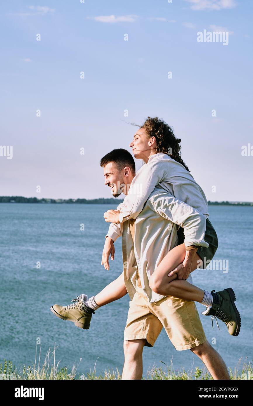 Fröhlich junge bärtige Mann geben Freundin Huckepack an der Küste während Sie haben Spaß während der Wanderung Stockfoto