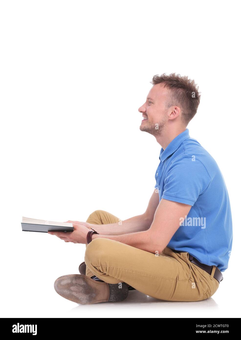 Seitenansicht eines lässigen jungen Mannes, der auf dem Boden sitzt und ein Buch hält, während er lächelt und vorwärts schaut, weg von der Kamera. Auf weißem Hintergrund Stockfoto
