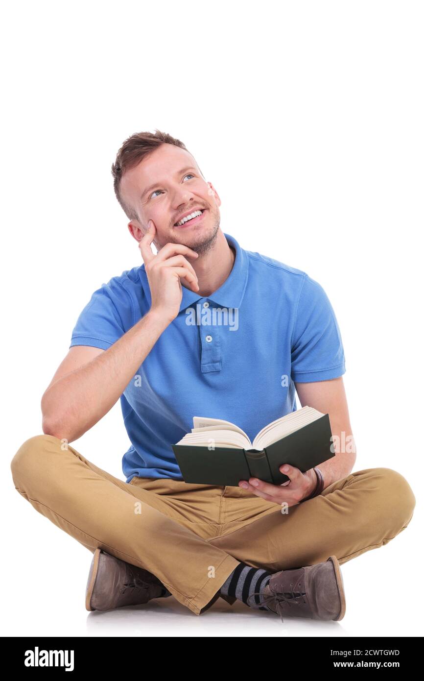 Lässiger junger Mann, der auf dem Boden sitzt, ein Buch hält und lächelnd von der Kamera wegschaut. Tagträumen. Auf weißem Hintergrund Stockfoto