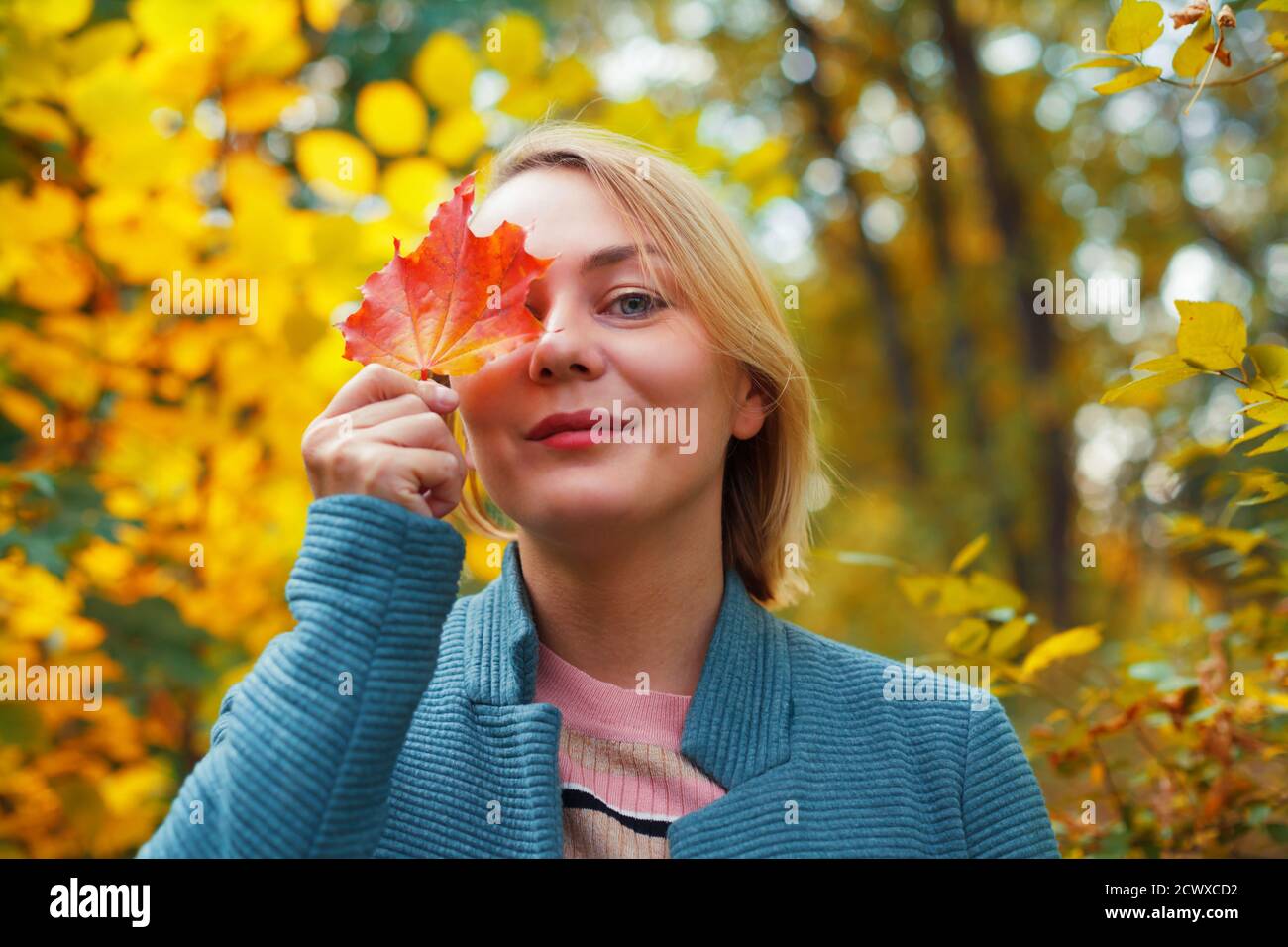 Lächelnde blonde Frau, die ein Auge mit rotem Ahornblatt bedeckt. Herbstkonzept. Im Freien Herbst weibliche Porträt Nahaufnahme Stockfoto