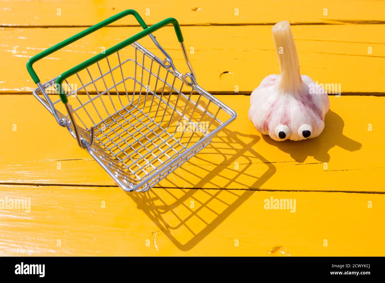 Lustiger Knoblauch mit Augen in der Nähe des kleinen Supermarktkorbs auf gelbem Hintergrund. Vegetarisches Konzept, Kopierraum Stockfoto