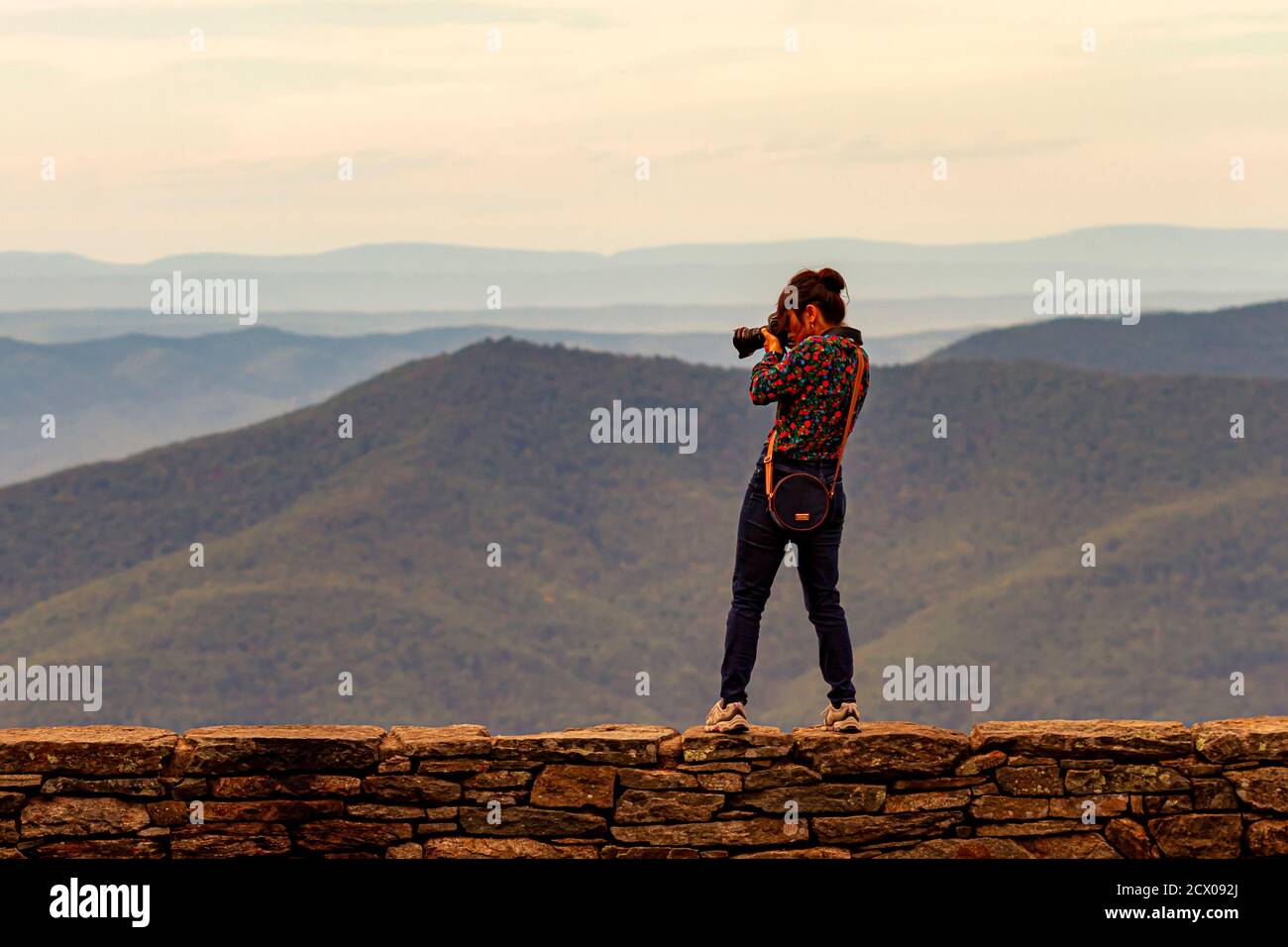 Ein junger kaukasischer Fotograf, der eine Seitentasche trägt, befindet sich auf einer Steinmauer an einer Klippe mit einem malerischen Blick auf die Skyline. Sie ist Taki Stockfoto
