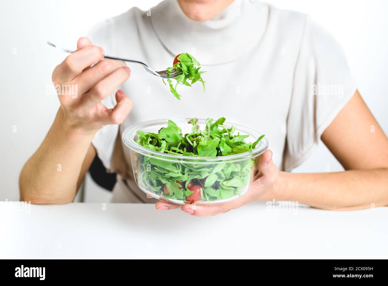 Unerkennbare junge Frau, die neben dem weißen Schreibtisch sitzt und einen Salat aus einer Glas-Lunchbox isst. Konzept der gesunden Ernährung Stockfoto