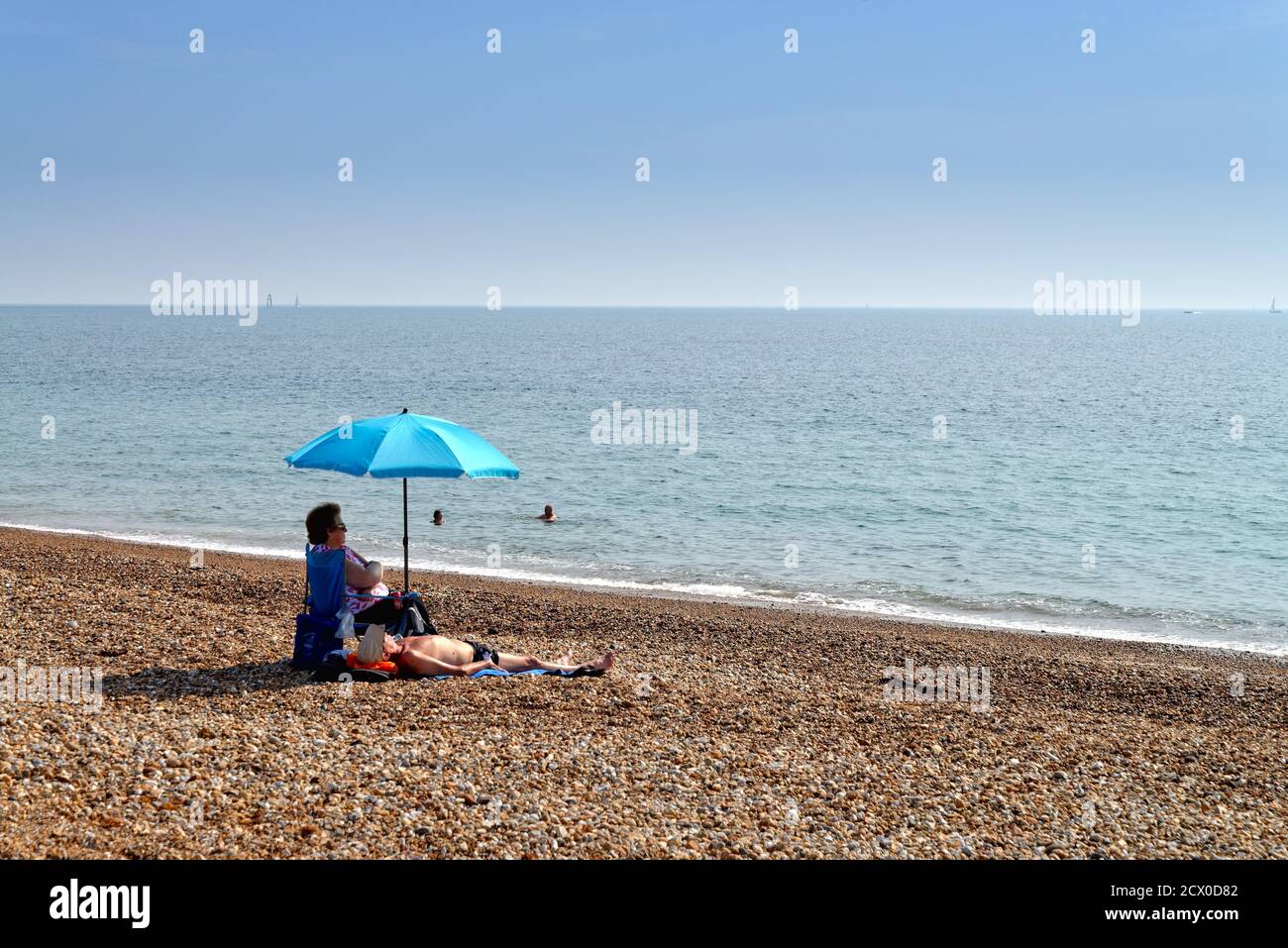Ein älteres Paar sitzt unter einem blauen Sonnenschirm am Strand von Hayling Island an einem heißen Sommertag, Hampshire England Stockfoto