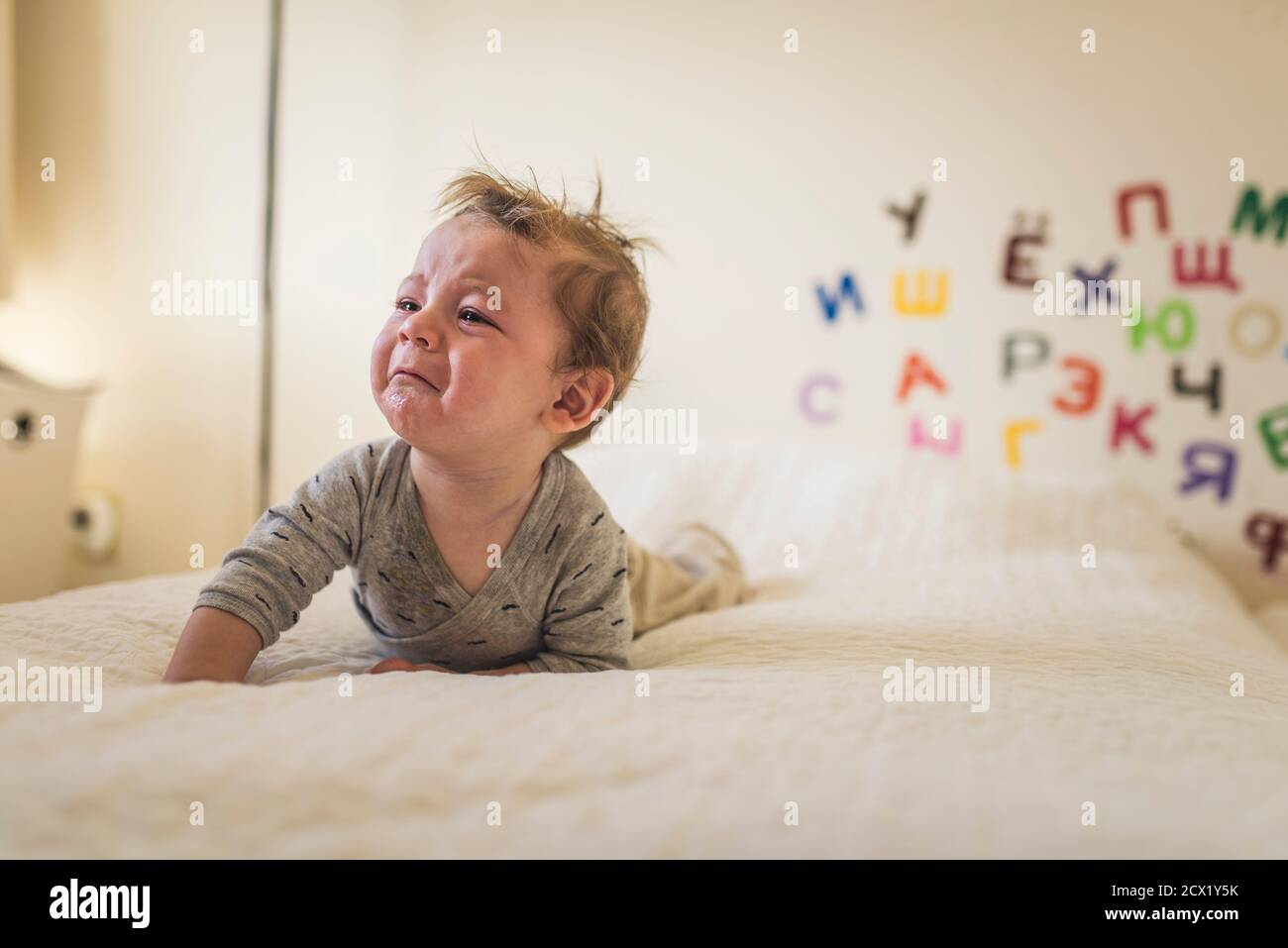 Weinendes Baby auf dem Bett mit weißer Decke und Alphabet auf Wand hinter ihm Stockfoto