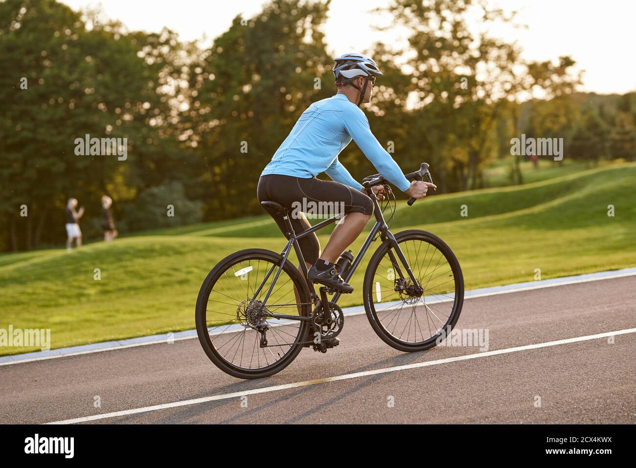 Radfahren im Freien. Seitenansicht des athletischen Mannes in Sportkleidung, der Fahrrad entlang einer Straße im Stadtpark bei Sonnenuntergang reitet, volle Länge. Gesunder aktiver Lebensstil und Sportkonzept Stockfoto
