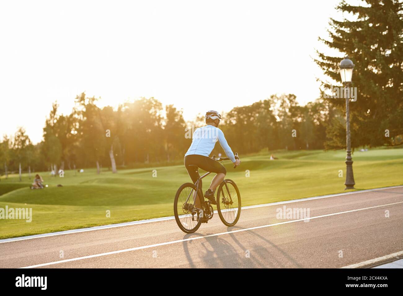 Perfekter Tag zum Radfahren. Rückansicht des athletischen Mannes in Sportkleidung, der Fahrrad entlang der Straße im Stadtpark bei Sonnenuntergang reitet, volle Länge. Gesunder aktiver Lebensstil und Sportkonzept Stockfoto