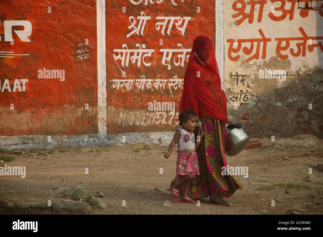 Indische Frau im roten Schleier mit Kind. Rajasthan, Indien. Dieses Bild enthält kulturell relevantes Material: Ein Sari ist ein südasiatisches weibliches Kleidungsstück, das aus einem Tuch mit einer Länge von 4 bis 8 Metern und einer Breite von 60 bis 1,20 Metern besteht, das typischerweise um die Taille gewickelt ist, wobei ein Ende über die Schulter drapiert ist und das Mittelriff entblößte. Der Sari wird normalerweise über einem Petticoat getragen. Der Sari ist mit Gnade verbunden und gilt weithin als Symbol der indischen Kultur. Stockfoto