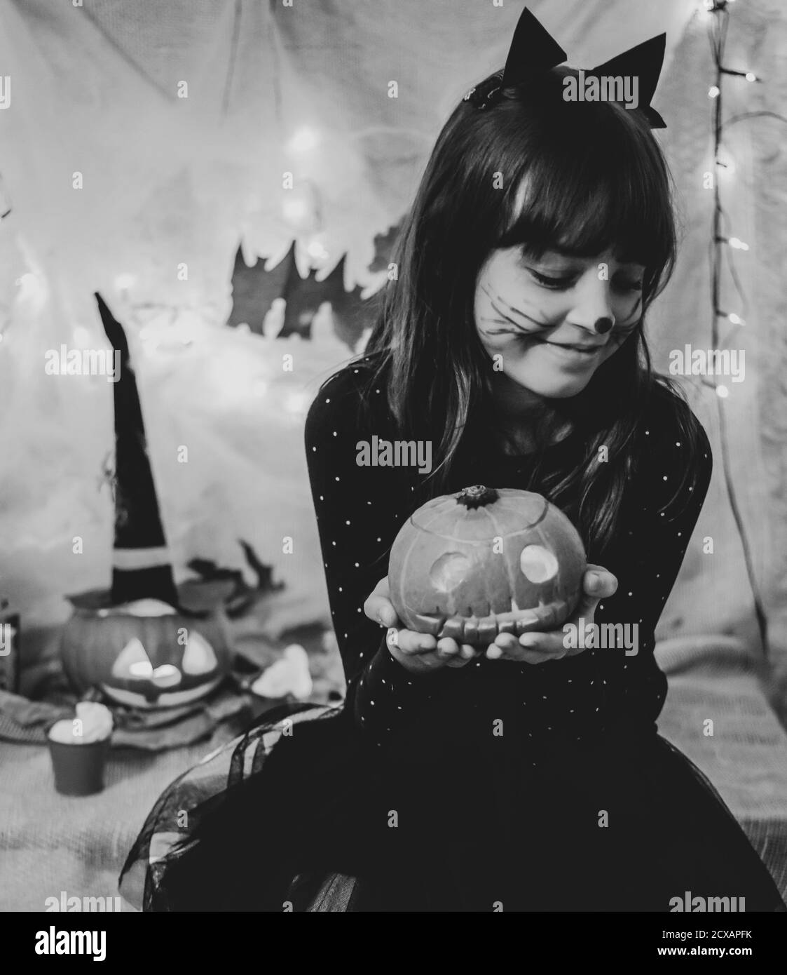 Halloween-Heimparty. Schwarz und weiß Foto von niedlichen lächelnden Mädchen in schwarz Katze Kostüm und geschnitzten Kürbis in den Händen Stockfoto