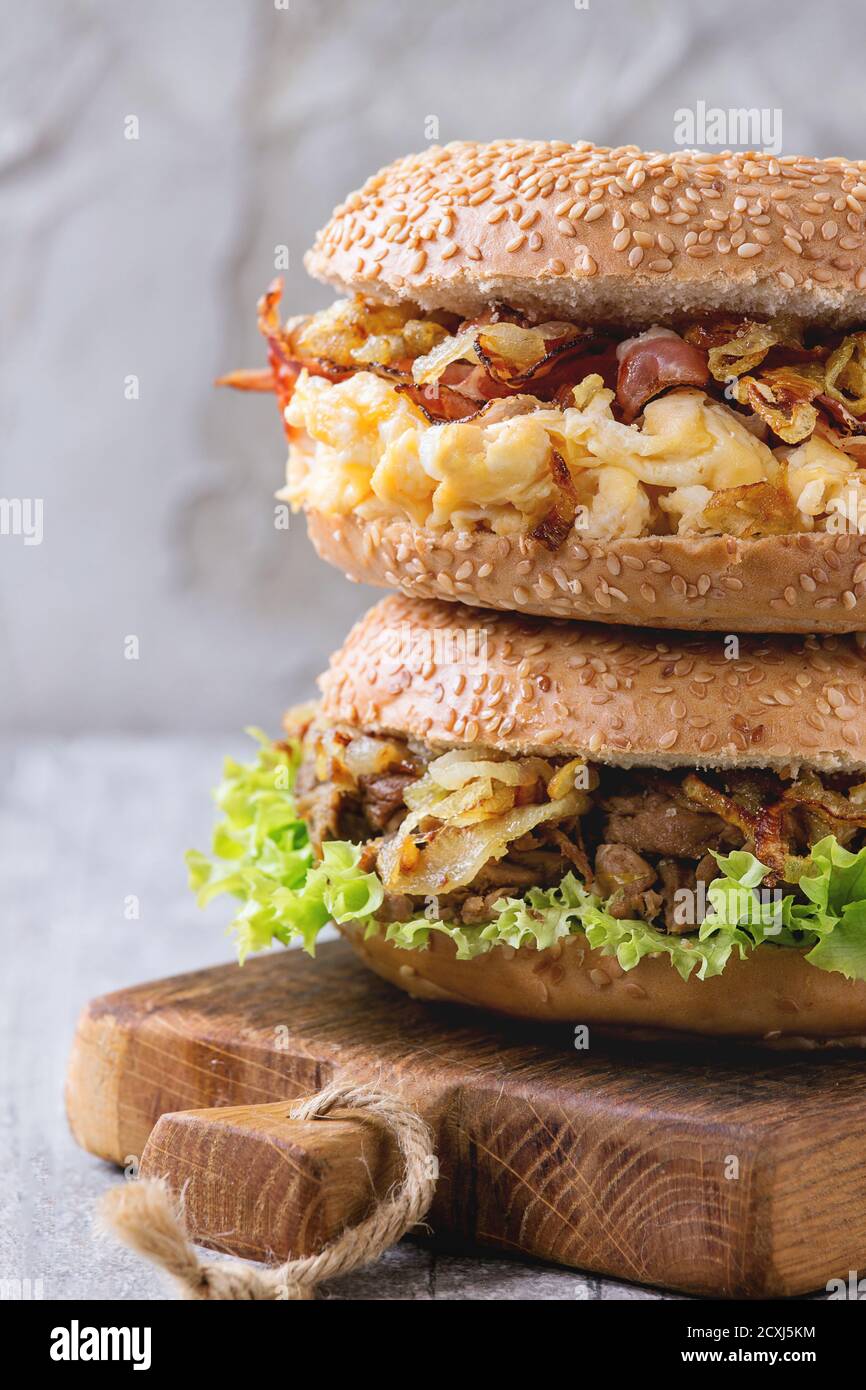 Stapel von zwei Bagels mit Eintopf Rindfleisch, Salat, Schinken, Rührei und gebratenen Zwiebeln auf kleinen hölzernen Schneidebrett über weiße Holztisch serviert. Stockfoto