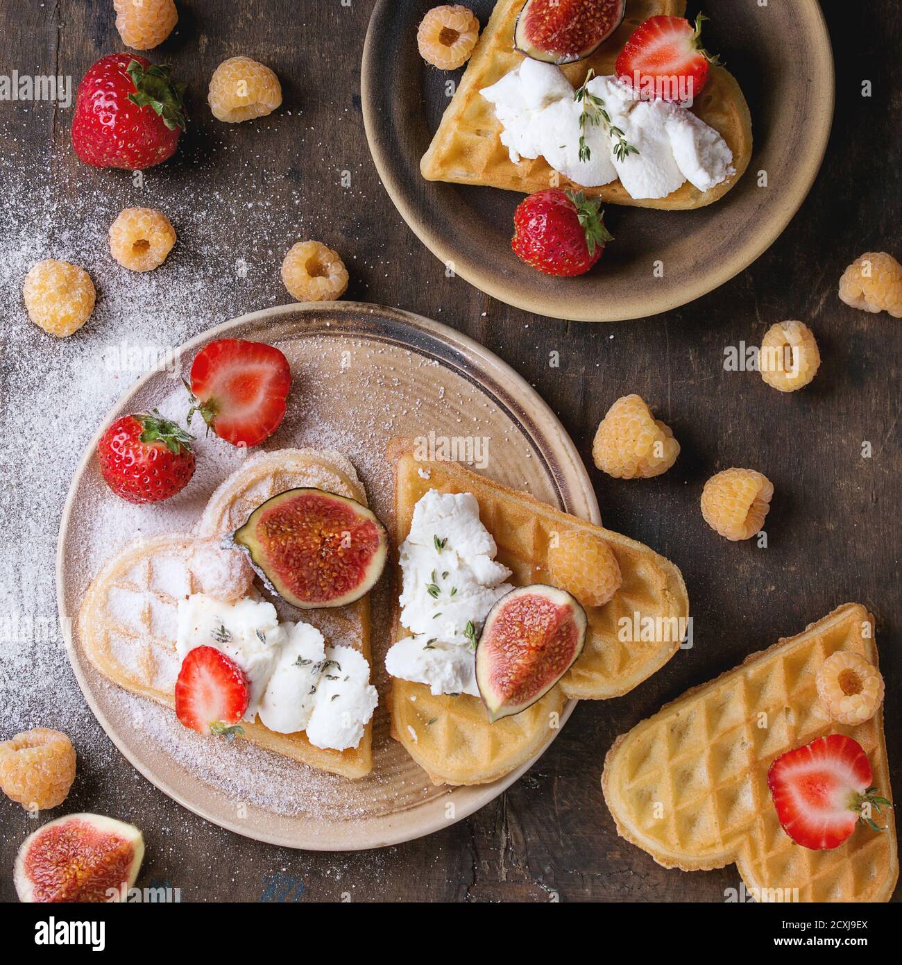 Waffeln als Herzform mit gelben Himbeeren, Erdbeeren, in Scheiben geschnittenen Feigen, Ricotta-Käse und Zuckerpulver auf Keramikplatten über dunklem Holzbackgroun Stockfoto