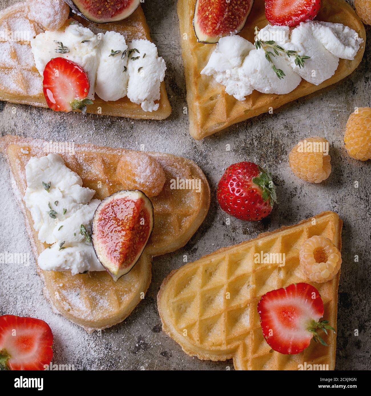 Waffeln als Herzform mit gelben Himbeeren, Erdbeeren, in Scheiben geschnittenen Feigen, Ricotta-Käse und Zuckerpulver über altem rostigen Metallhintergrund. Draufsicht. Stockfoto