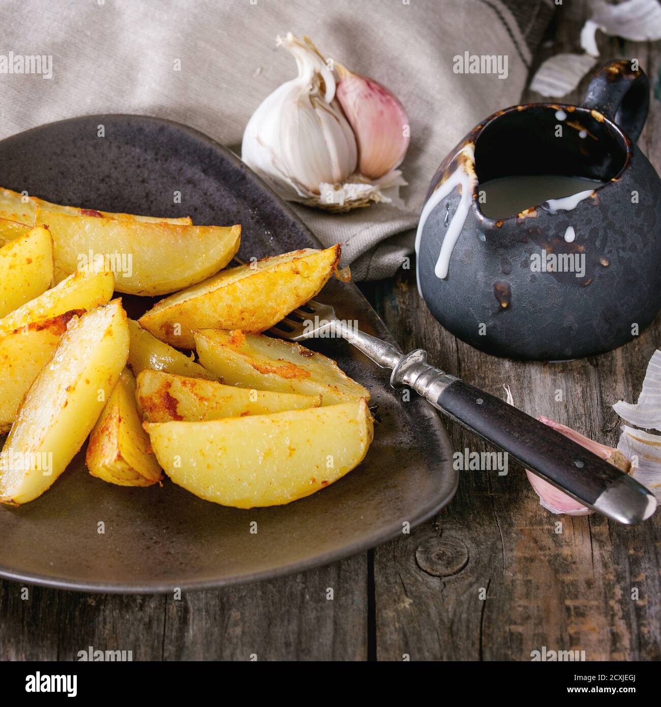 Gebratene Landkartoffeln auf brauner Keramikplatte, serviert mit hausgemachter Aioli-Sauce in Krug, Knoblauch und Vintage-Gabel auf grauer Leinenserviette über Holzrücken Stockfoto