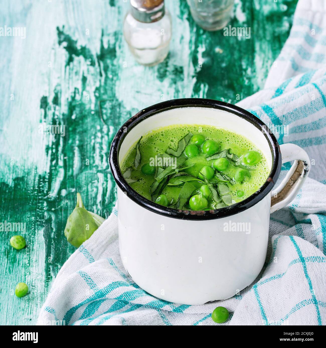 White vintage Becher mit Erbsen Cremesuppe mit geschnittenem Basilikum, serviert mit Salz und Pfefferstreuer auf weißen Küche Handtuch über grünen und weißen Holzmöbeln backgr Stockfoto