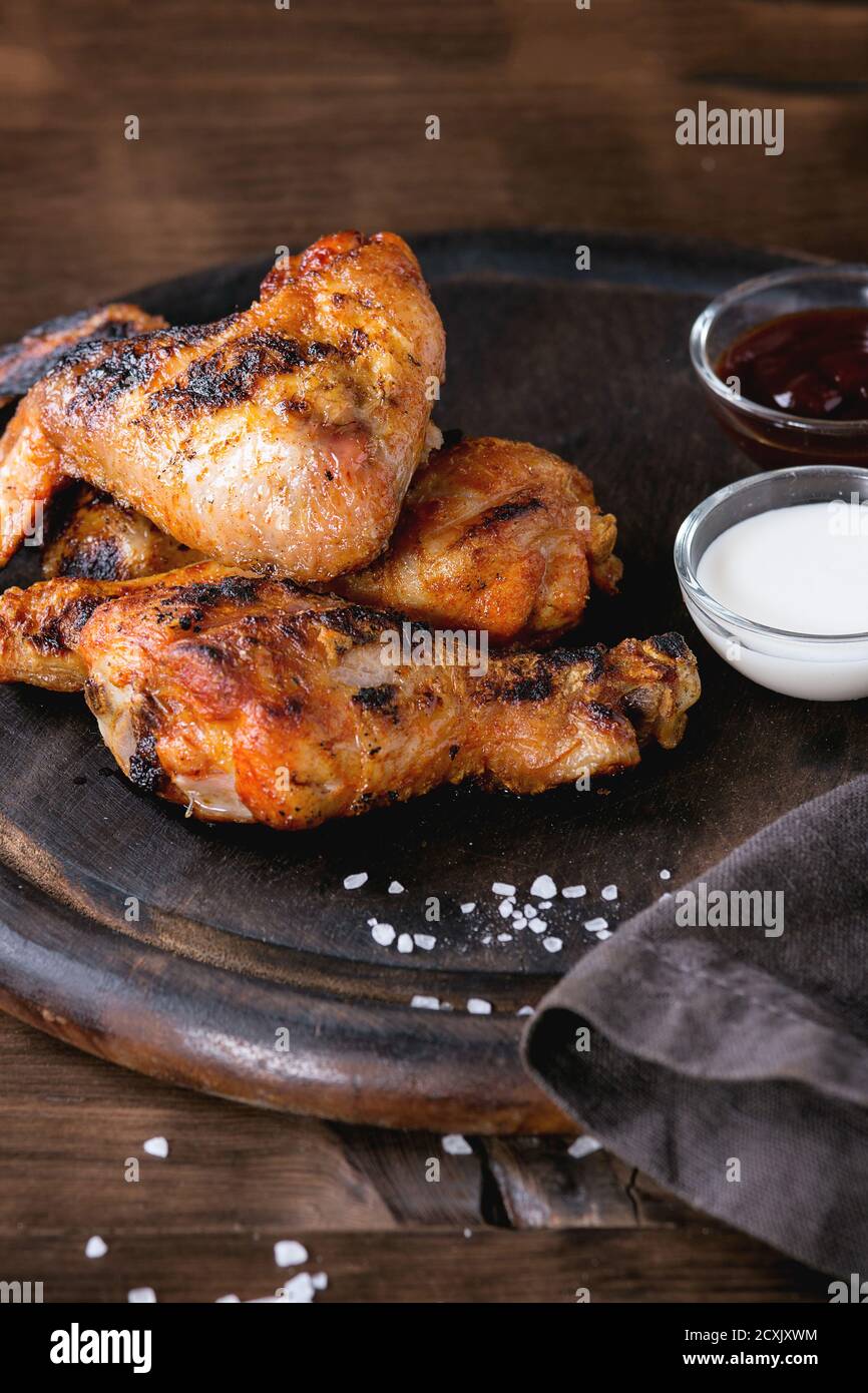 Gegrilltes BBQ Hähnchenfleisch Flügel und Beine serviert auf runden Holz Schneidebrett mit zwei Saucen und Salz auf grauem Textil auf dunklem Holzhintergrund. Stockfoto
