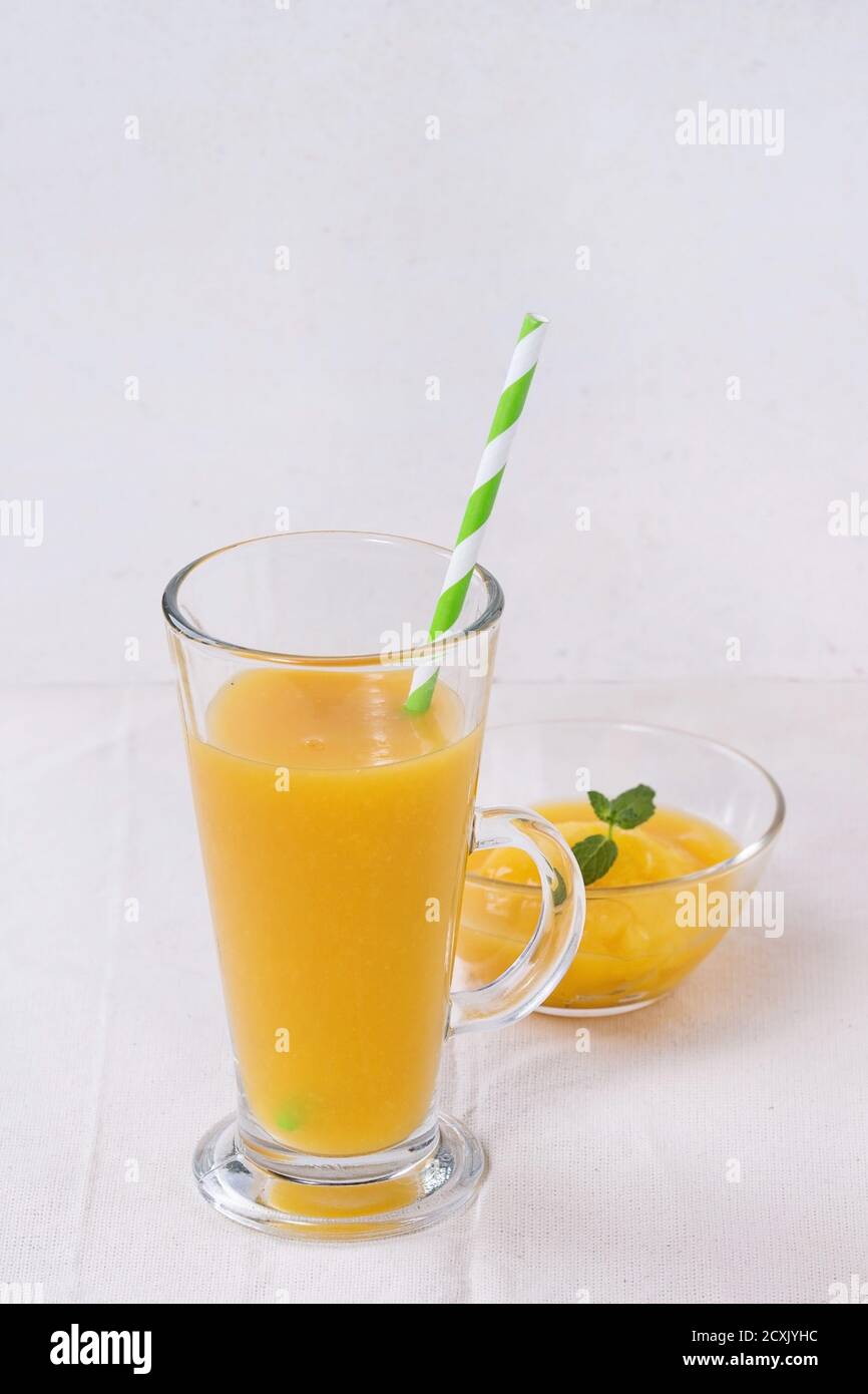 Ein Glas frischer Mango-Smoothie mit gestreiftem grünem Cocktailröhrchen und ein Mango-Dessert mit Minze, serviert auf einer weißen Tischdecke mit weißer strukturierter Ba Stockfoto