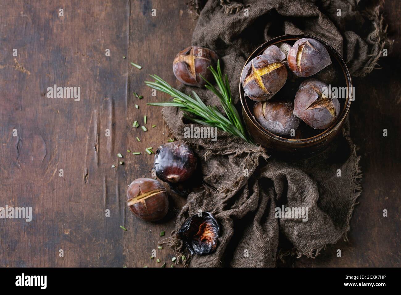 Gebratene Kastanien in der Asche mit Rosmarin in Blechdose auf Sackleinen über alten dunklen hölzernen Hintergrund. Draufsicht mit Platz für Text. Stockfoto