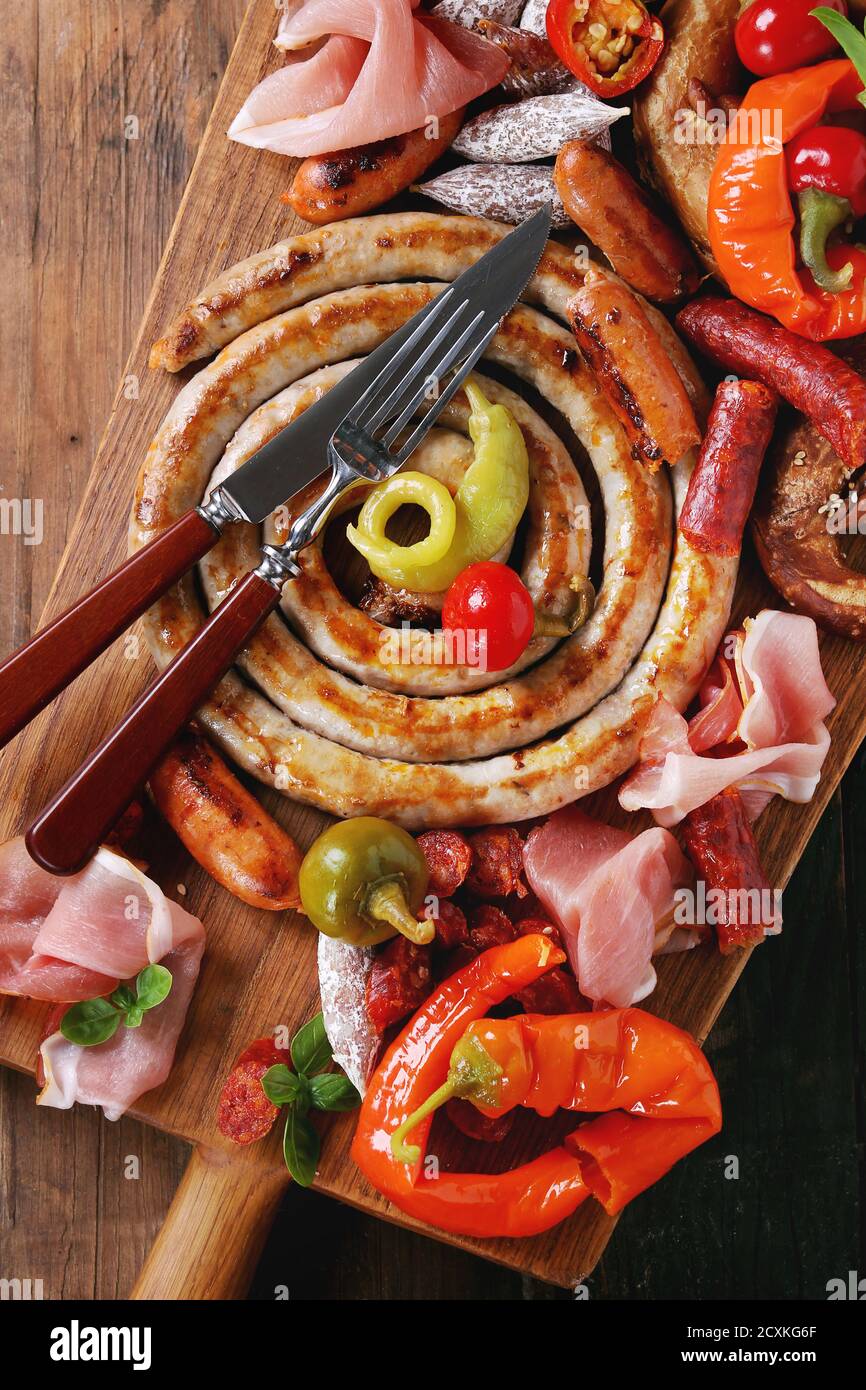 Auswahl an Fleischsnacks Bratwürste, Wienerwurst, Schinken, marinierte Chilischoten in gesalzenen Brezeln mit Besteck auf Holzhackbrett über da serviert Stockfoto