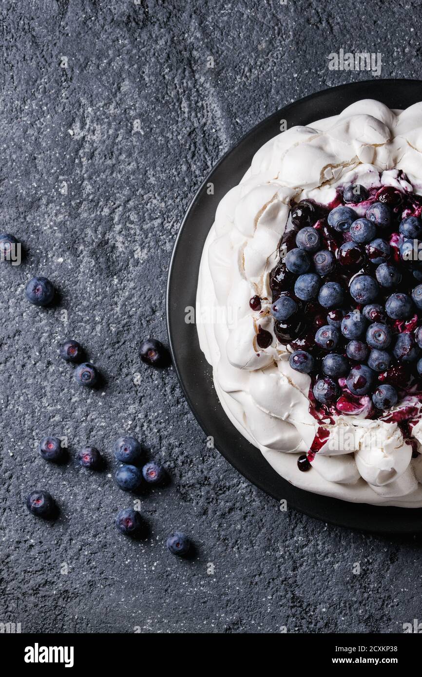 Hausgemachtes Baiser Kuchen Pavlova mit Schlagsahne, frische Heidelbeeren und Blaubeerensauce auf Vintage Kuchen stehen auf schwarze Betontextur Hintergrund. T Stockfoto