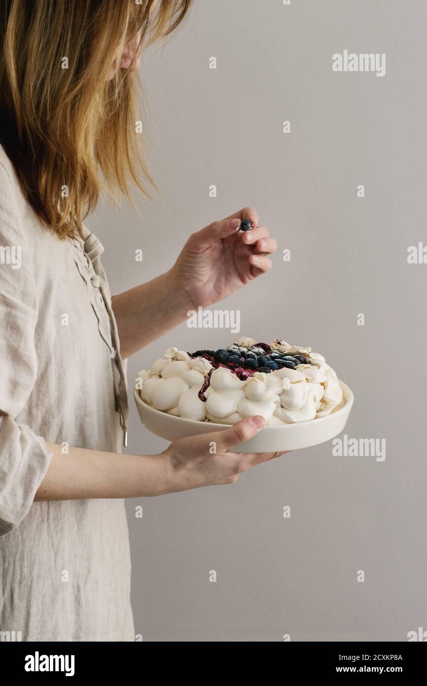 Frau Kuchen schmecken. Hausgemachtes Baiser Kuchen Pavlova mit Schlagsahne, Puderzucker, frische Heidelbeeren und Blaubeerensauce in weiblichen Händen über grau w Stockfoto