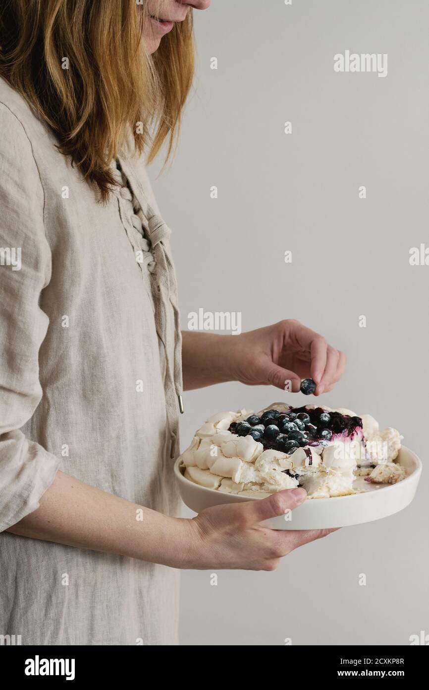 Frau Kuchen schmecken. Hausgemachtes Baiser Kuchen Pavlova mit Schlagsahne, Puderzucker, frische Heidelbeeren und Blaubeerensauce in weiblichen Händen über grau w Stockfoto
