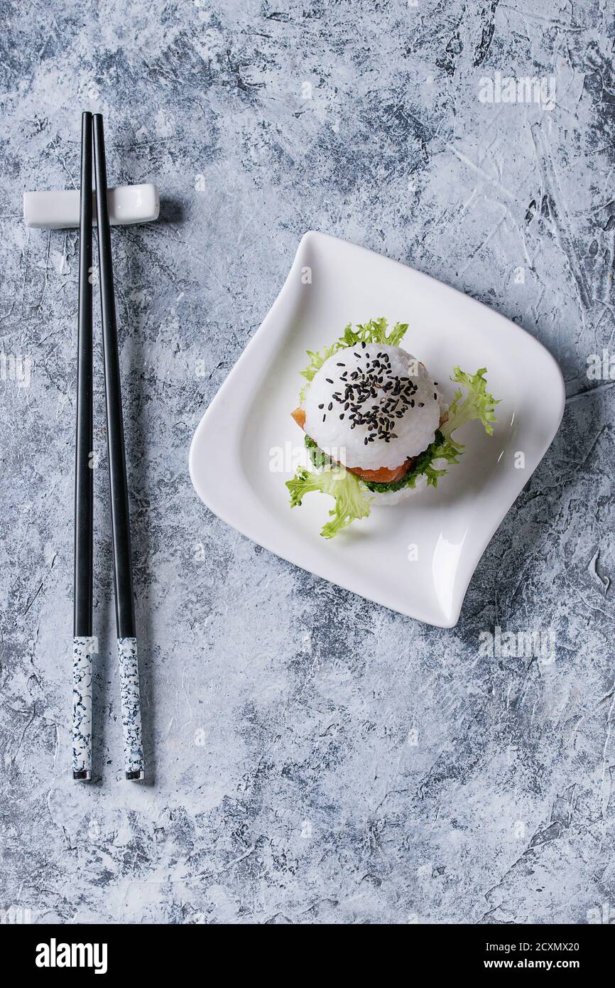Mini Reis Sushi Burger mit geräuchertem Lachs, grünem Salat und Saucen, schwarzer Sesam serviert auf weißem quadratischen Teller mit Essstäbchen über grauem Beton Backgr Stockfoto