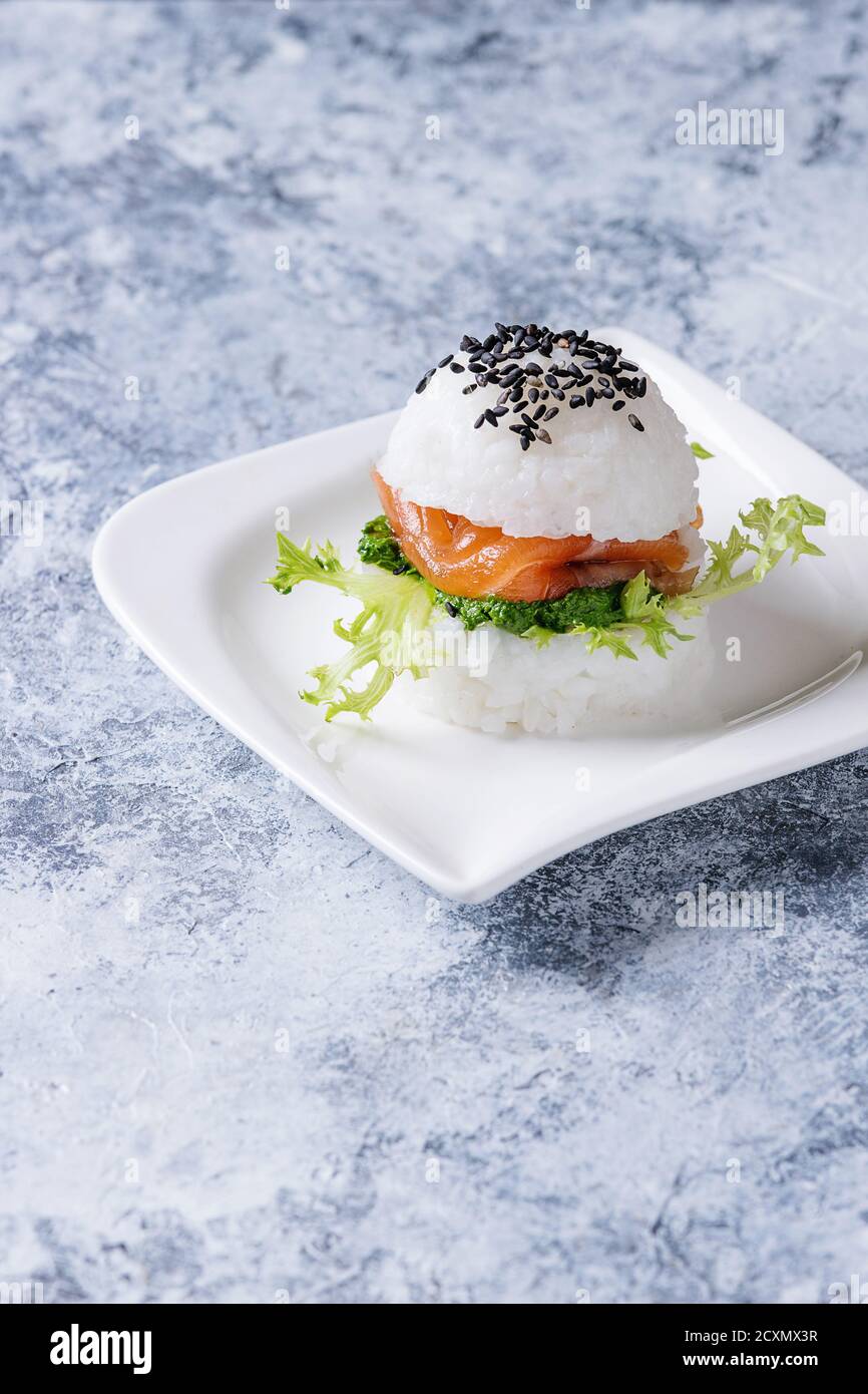 Mini-Reis-Sushi-Burger mit geräuchertem Lachs, grünem Salat und Saucen, schwarzem Sesam serviert auf weißem quadratischem Teller auf grauem Betonboden. Moderne hea Stockfoto