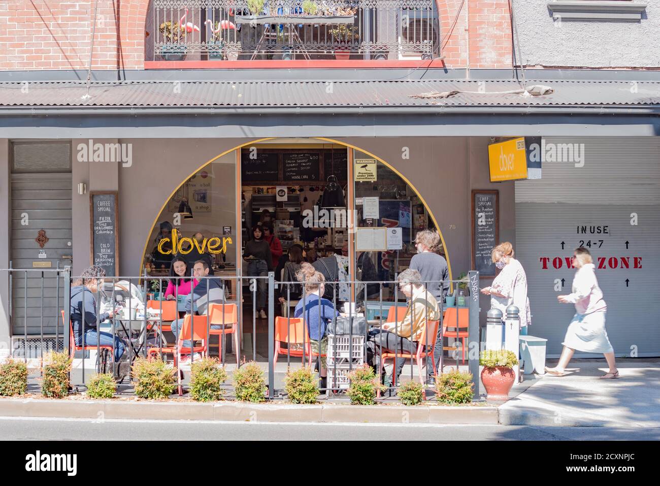Vor einem Café sitzen Menschen in geselligem Abstand, während eine Frau mit Maske im Vorort Annandale in Sydney, New South Wales, Australien, vorbeiläuft Stockfoto