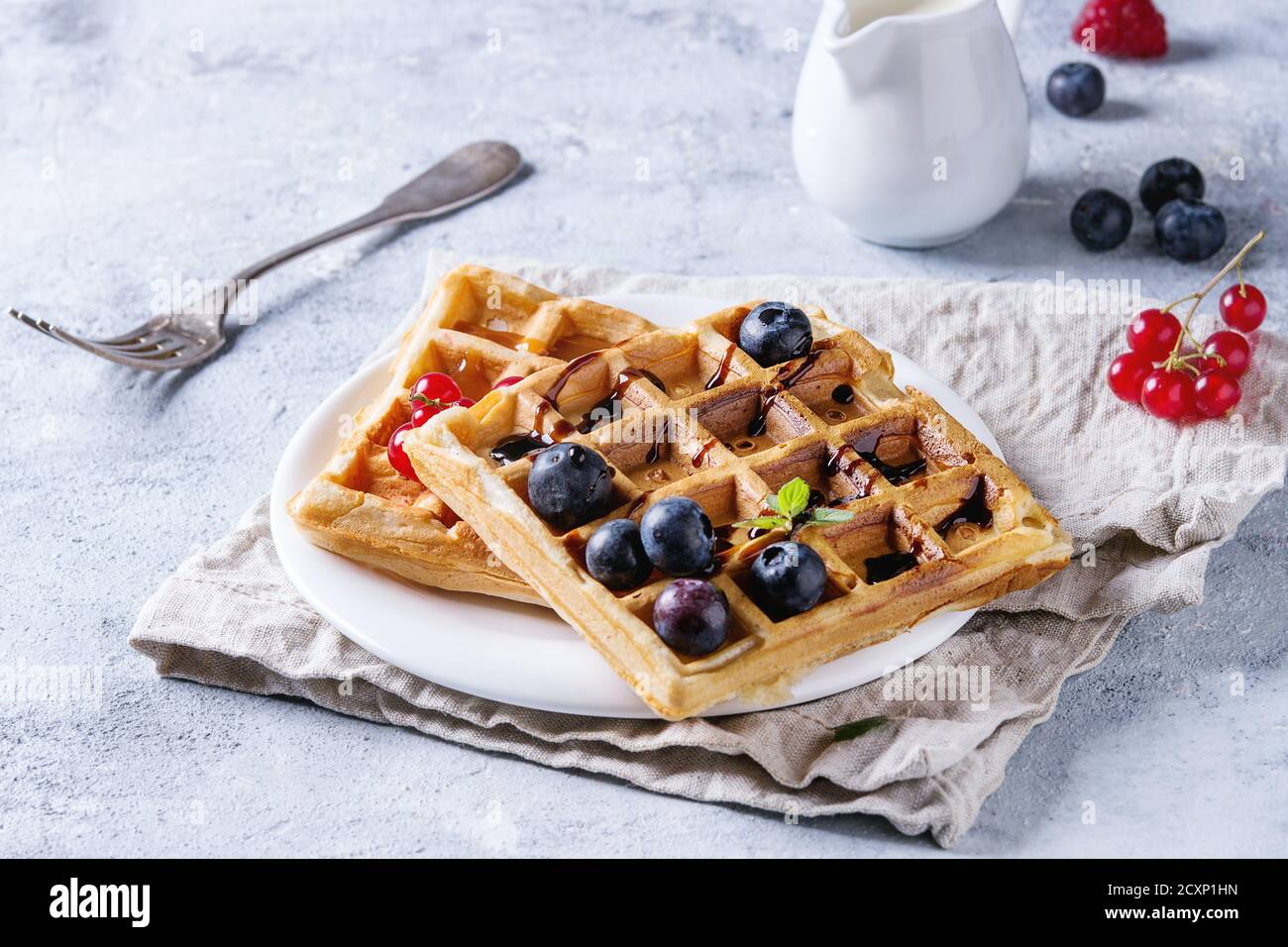 Frühstück mit hausgemachten quadratischen belgischen Waffeln mit frischen reifen Beeren Heidelbeere, Himbeere, rote Johannisbeere, Balsamico-Sauce auf weißem Teller mit Krug mi Stockfoto